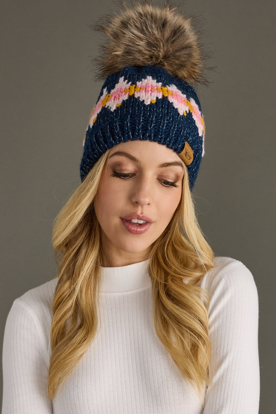 Woman wearing a blue knit beanie with a colorful pattern and fur pom-pom on a gray background