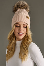 Woman wearing a blush knit beanie with a fur pom-pom against a gray background