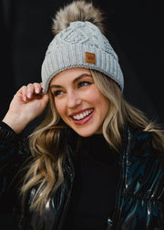 A woman with long blonde hair smiles while looking slightly to the side, wearing a light gray knit beanie with a faux-fur pom-pom and a small leather patch on the front. She’s dressed in a dark, glossy winter jacket, posed against a plain dark background that highlights her warm expression and cozy cold-weather style.
