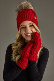 A smiling woman models the Panache Apparel Collection Red Cable Knit Pom Hat, featuring a cozy fleece lining and a stylish faux fur pom, paired with matching red knit mittens and a black long-sleeve top against a plain grey background.