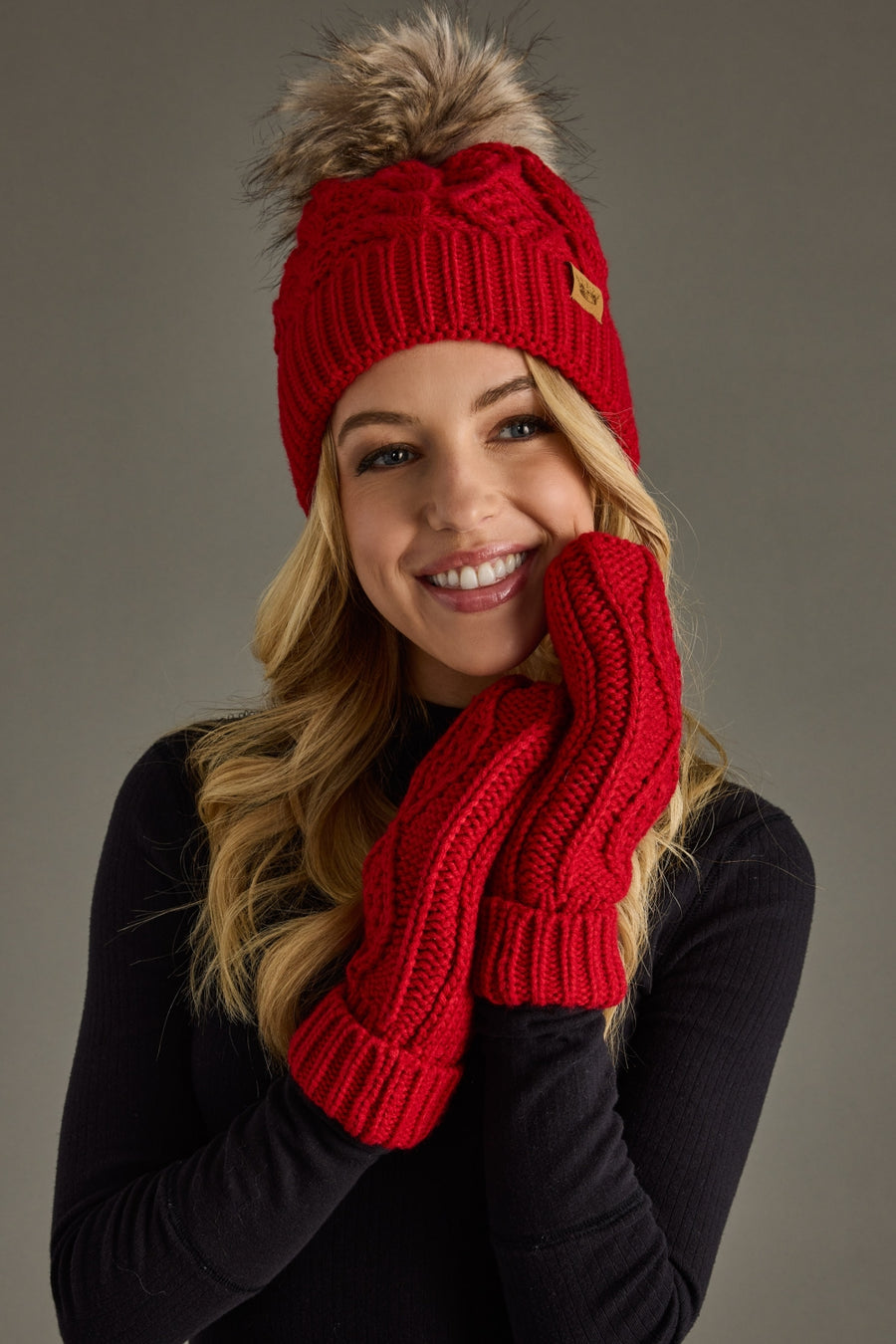 A smiling woman models the Panache Apparel Collection Red Cable Knit Pom Hat, featuring a cozy fleece lining and a stylish faux fur pom, paired with matching red knit mittens and a black long-sleeve top against a plain grey background.
