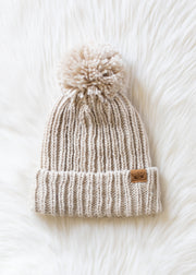 Beige knit beanie with pom-pom on a white textured background
