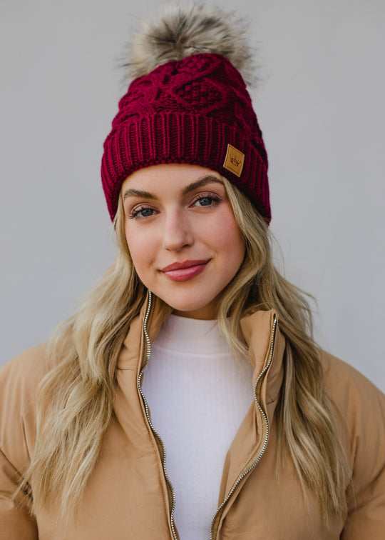 Woman wearing a red knit beanie with a pom-pom and a beige jacket against a plain background