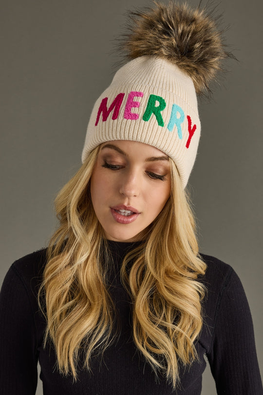 Woman wearing a cream beanie with 'MERRY' in colorful letters and a fur pom-pom on a gray background