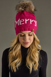 A woman with long blonde hair wears the Panache Apparel Collection Merry Pom Hat in magenta, fleece lined for warmth and featuring "MERRY" in white, paired with a black turtleneck against a plain gray background.