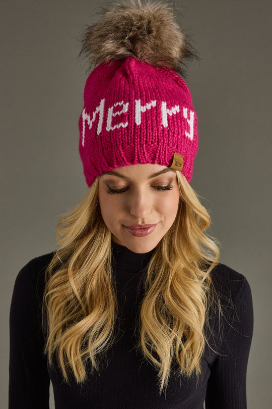 A woman with long blonde hair wears the Panache Apparel Collection Merry Pom Hat in magenta, fleece lined for warmth and featuring "MERRY" in white, paired with a black turtleneck against a plain gray background.