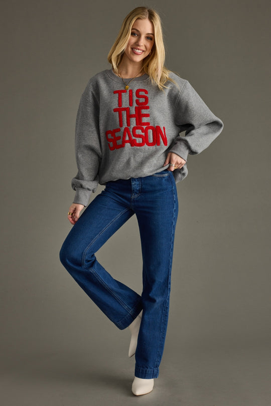 Woman wearing a gray sweater with 'TIS THE SEASON' text and blue jeans on a gray background