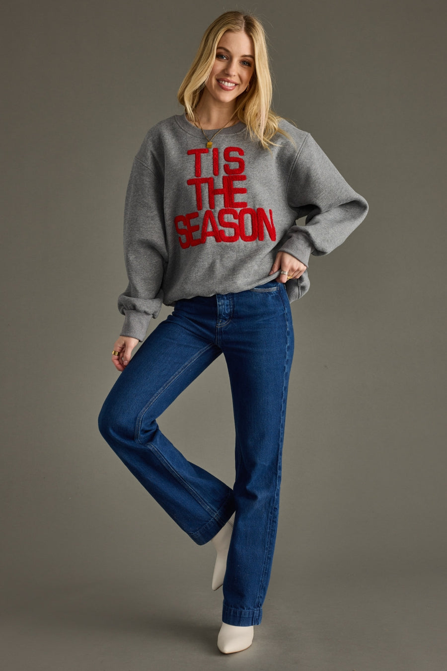 Woman wearing a gray sweater with 'TIS THE SEASON' text and blue jeans on a gray background
