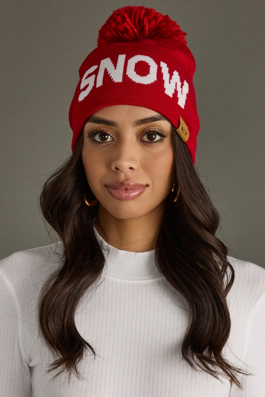 A woman with long, wavy dark hair wears the Panache Apparel Collection Snow Pom Hat - Red, featuring a white "SNOW" design and pom-pom, paired with a white ribbed turtleneck sweater against a plain gray background.