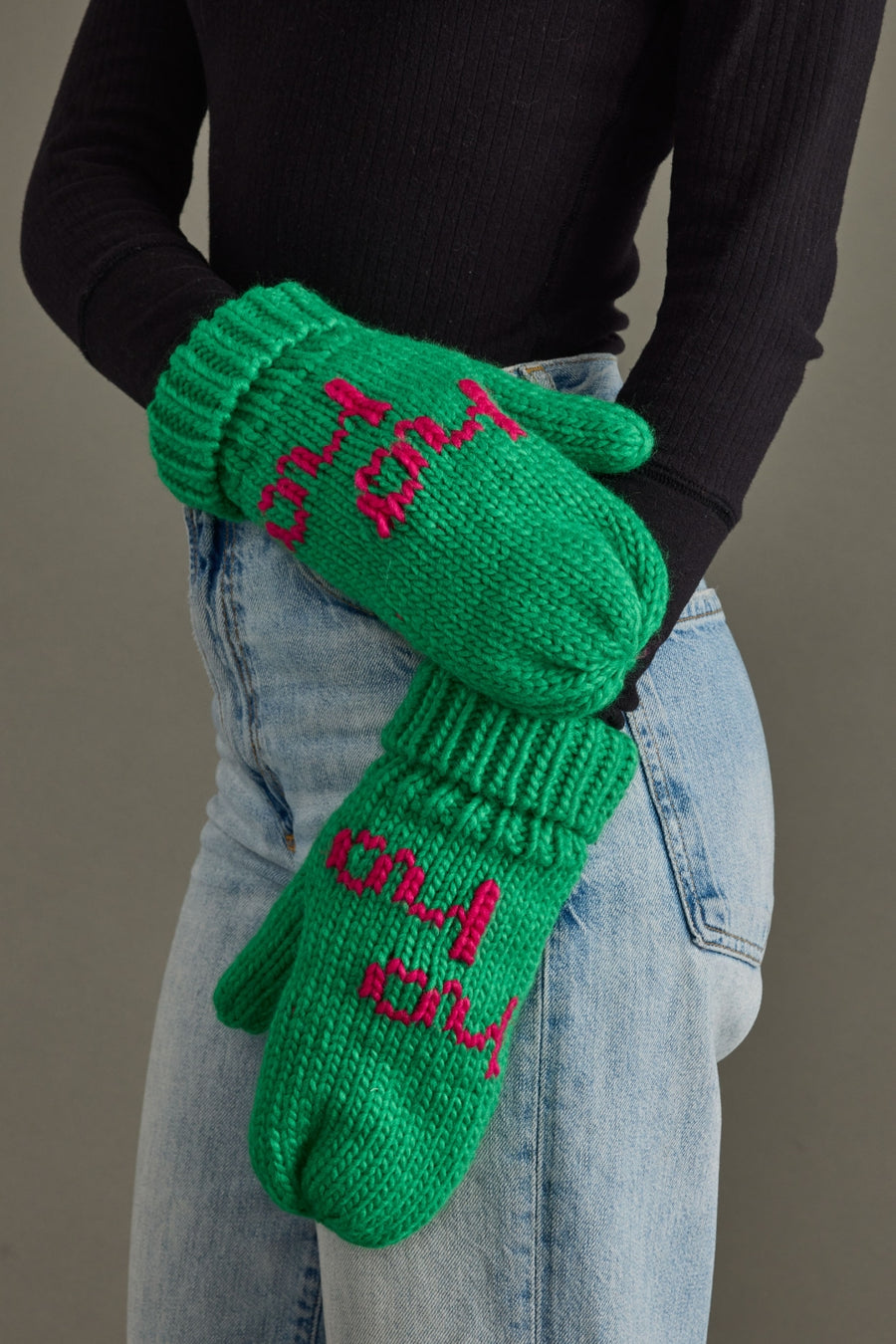 Green mittens with pink text worn by a person against a neutral background
