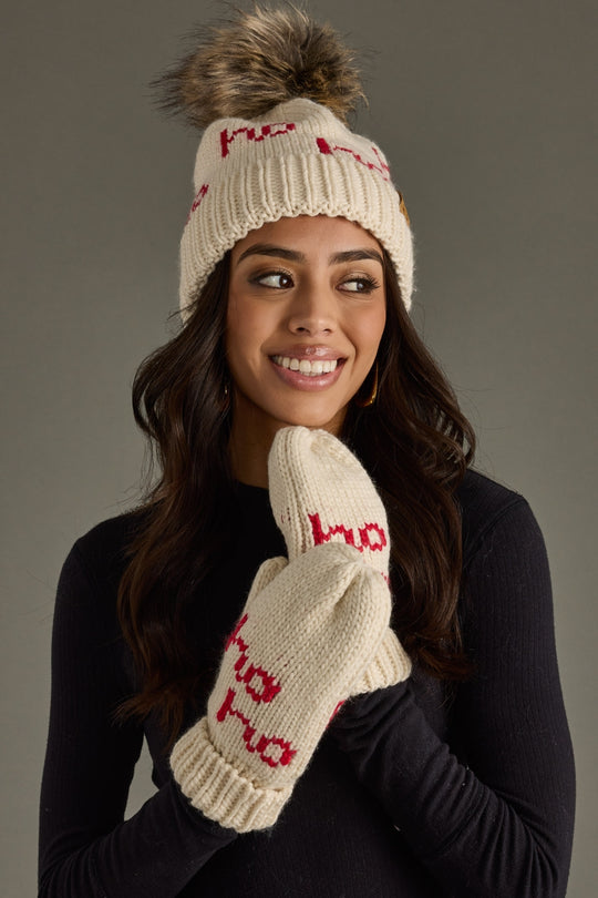 Woman wearing a cream knit hat with red text and matching mittens on a gray background
