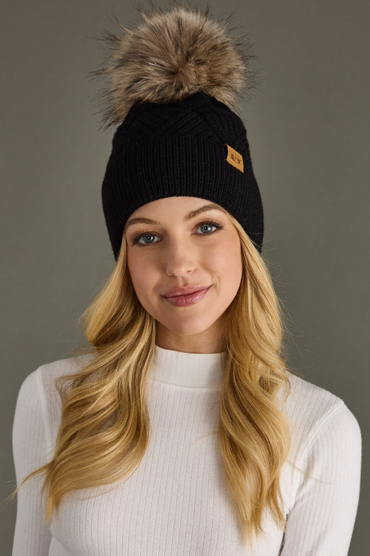 Woman wearing a black beanie with a fur pom-pom against a gray background