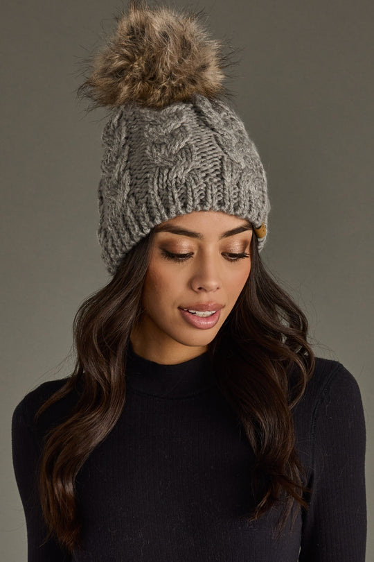 Woman wearing a gray knit beanie with a pom-pom against a gray background