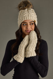 Woman wearing a cream knit beanie with a pom-pom and matching gloves against a gray background
