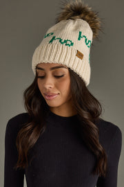 Woman wearing a cream beanie with green text and a fur pom-pom on a gray background
