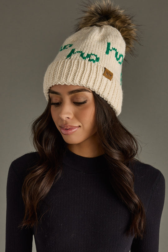 Woman wearing a cream beanie with green text and a fur pom-pom on a gray background