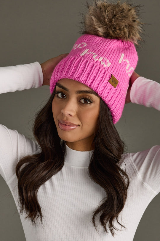 A woman adjusts the Panache Apparel Collection Ho Ho Ho Pom Hat - Pink & White, a bright pink knit beanie with fleece lining, white pattern, and faux fur pom-pom, paired with a white long-sleeve top against a neutral background.