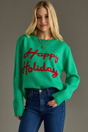 A woman with long blonde hair wears the Panache Apparel Collection Happy Holidays Sweater - Green, featuring red text, paired with blue jeans. She smiles slightly while standing against a plain gray background.