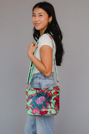 A woman with long dark hair, dressed in a white t-shirt and blue jeans, stands sideways and smiles while holding the Panache Apparel Collection Libby Floral Crossbody against a plain gray background.