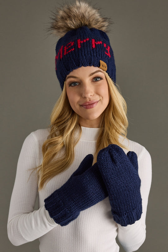 Woman wearing a navy knit hat with red text and fur pom-pom, paired with matching mittens on a gray background.
