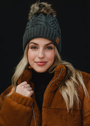 A woman with long blonde hair looks directly at the camera with a soft, relaxed expression, wearing a dark gray cable-knit beanie with a faux-fur pom-pom and a small leather logo patch on the front. She’s styled in a black top layered under a warm brown jacket, set against a clean, dark background that emphasizes her natural makeup and cozy fall–winter aesthetic.