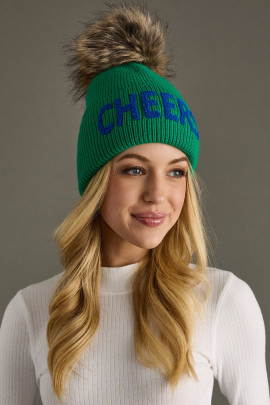 Woman wearing a green beanie with 'CHEERS' text and fur pom-pom on a gray background