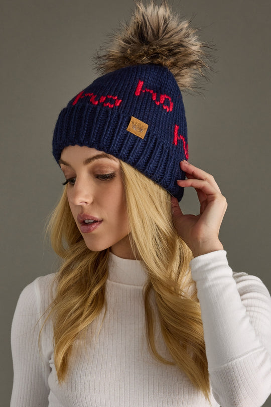 Woman wearing a navy blue beanie with red text and a fur pom-pom, against a neutral background.