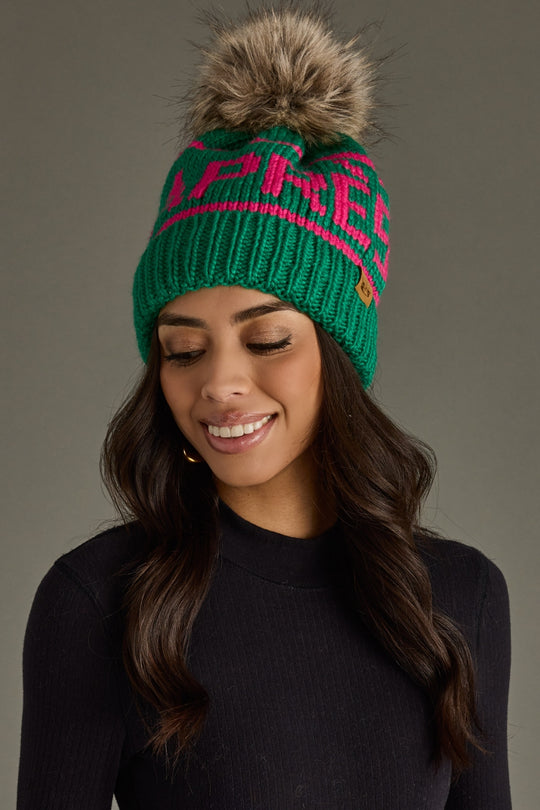 Woman wearing a green knit beanie with pink text and a fur pom-pom on a gray background