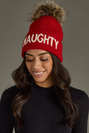 Woman wearing a red 'NAUGHTY' beanie with a fur pom-pom on a gray background