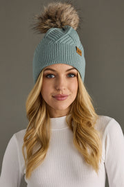 Woman wearing a teal knit beanie with a fur pom-pom against a gray background