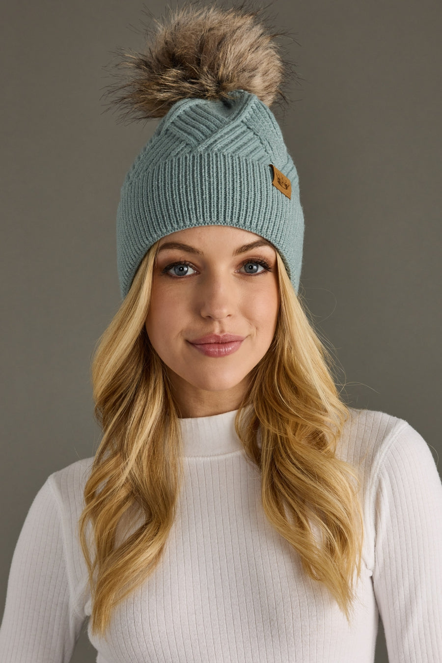 Woman wearing a teal knit beanie with a fur pom-pom against a gray background