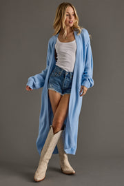 Woman wearing a long blue cardigan, white top, denim shorts, and beige boots on a gray background