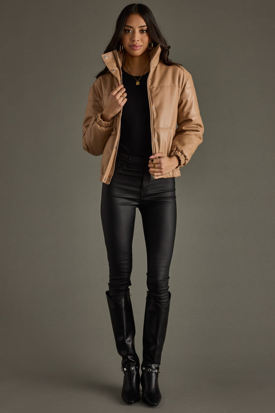 Woman wearing a tan jacket, black top, and black leather pants against a gray background