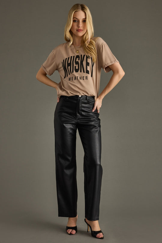 A woman poses against a gray background, wearing the Panache Apparel Collection’s Light Brown Whiskey Weather Tee in an oversized fit, paired with black leather pants and open-toe heels. She smiles slightly with one hand on her hip.