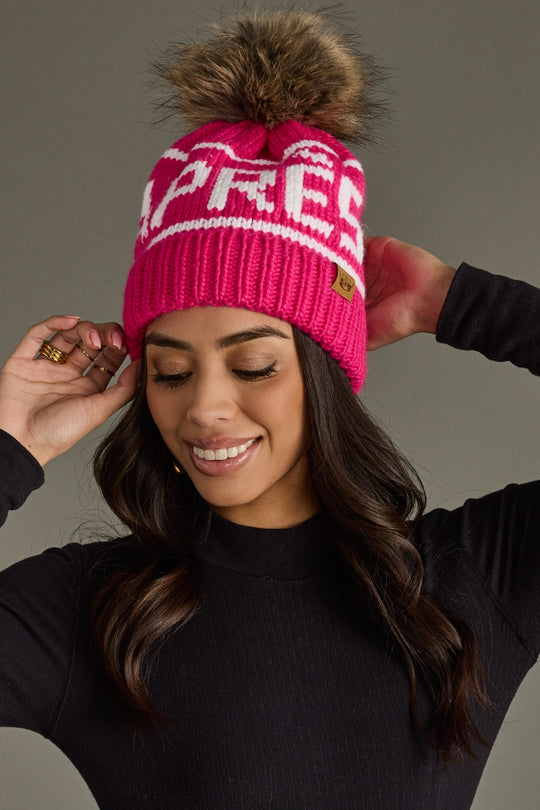 Woman wearing a pink beanie with a fur pom-pom against a gray background