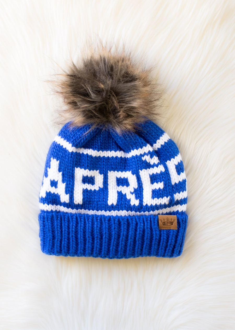 Blue knit beanie with white text and fur pom-pom on a white background