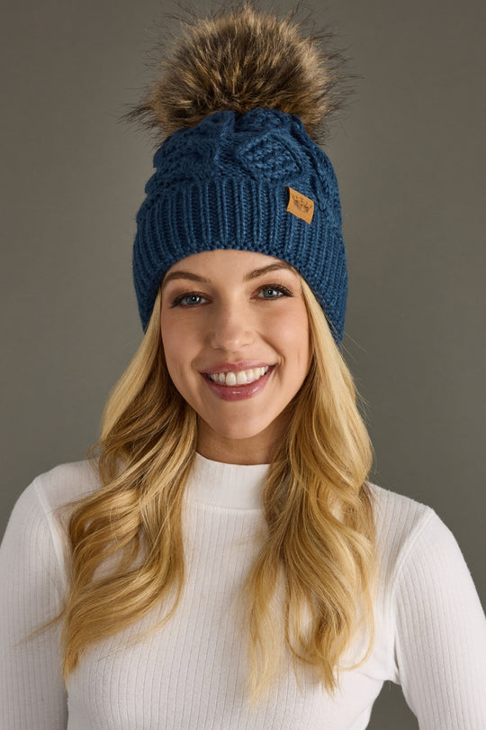 Woman wearing a blue knitted beanie with a fur pom-pom against a gray background