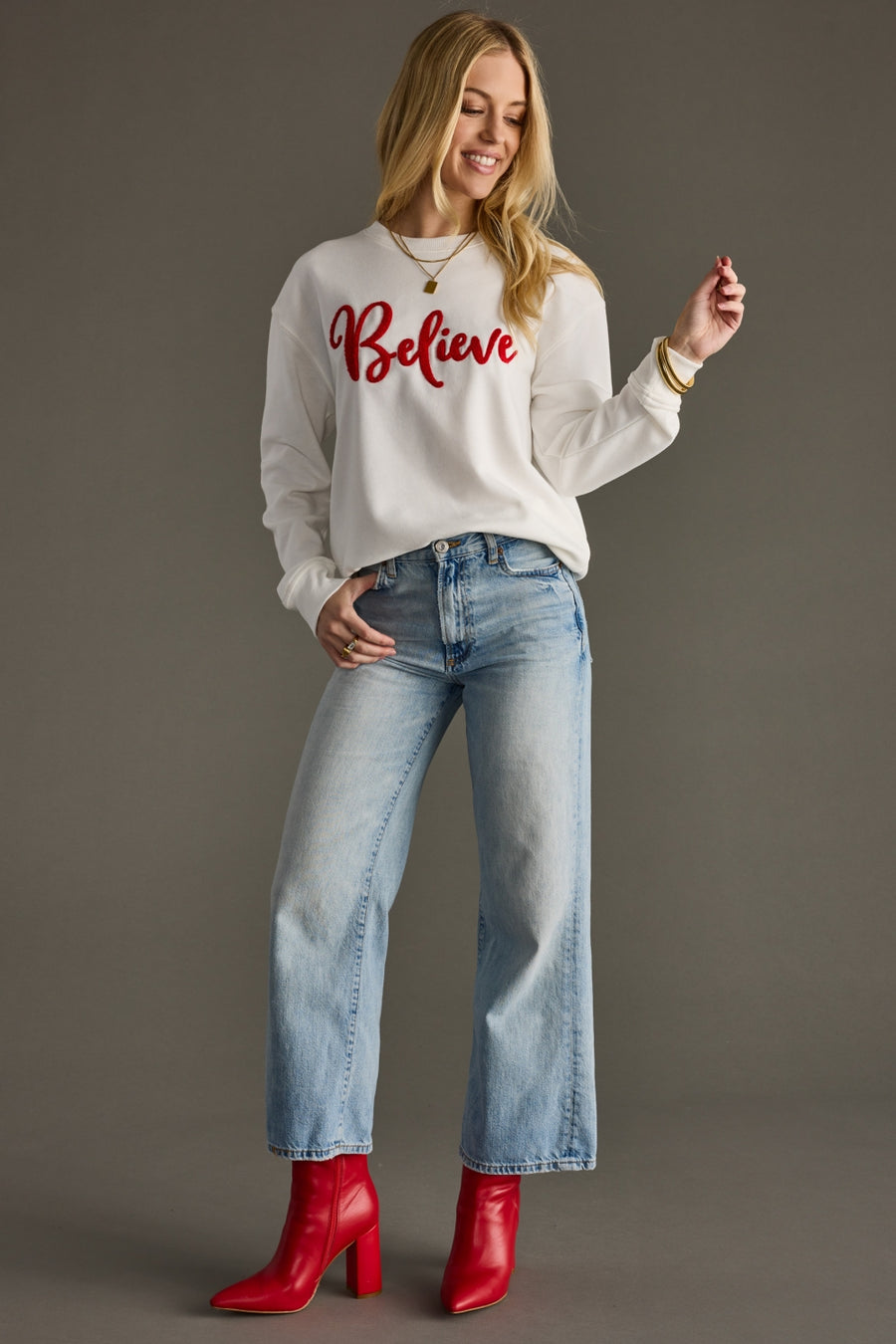 Woman wearing a white sweatshirt with 'Believe' text, blue jeans, and red boots on a gray background