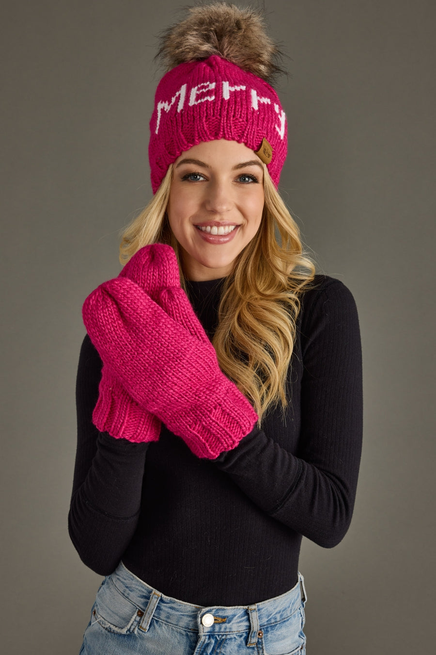 Woman wearing a pink 'Merry' beanie and matching gloves against a gray background