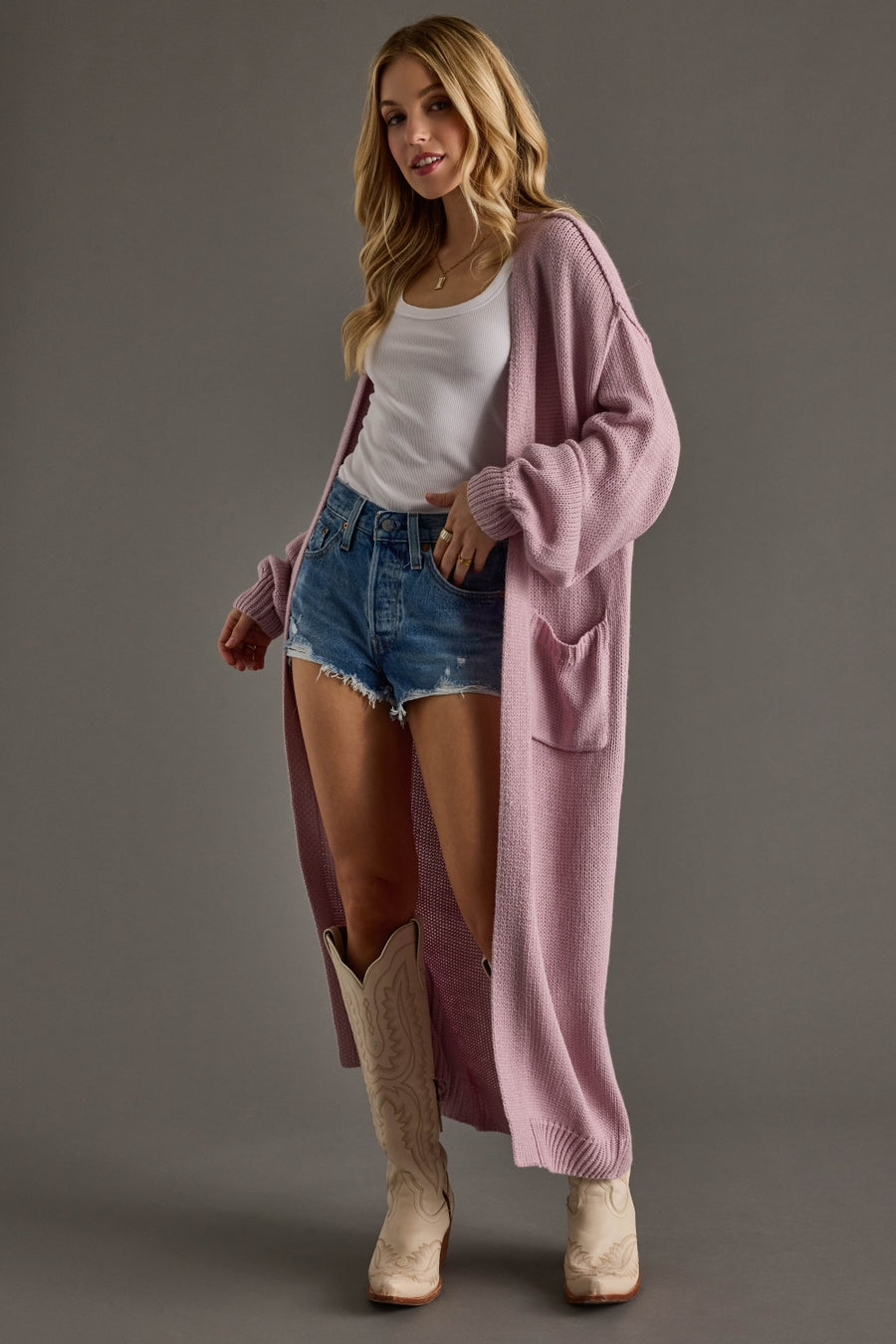 Woman wearing a long pink cardigan, white top, denim shorts, and beige boots on a gray background