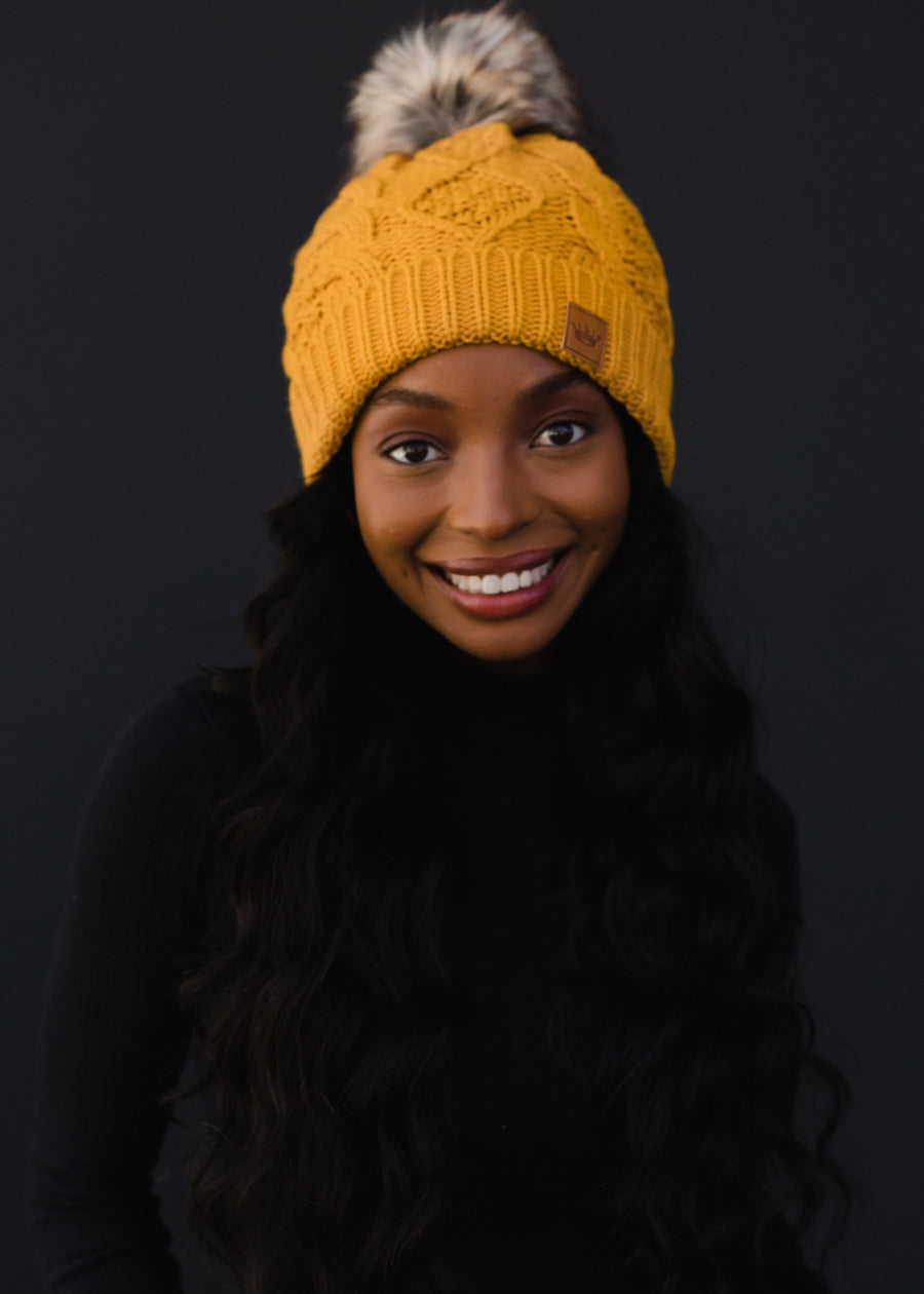 Woman wearing a yellow knit beanie with a fur pom-pom against a black background