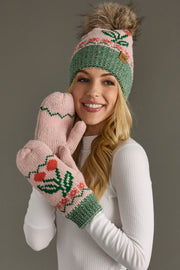 A smiling woman in a white sweater wears the Panache Apparel Collection Light Pink Cherry Pattern Pom Hat, a pink and green fleece-lined knit hat with a faux fur pom-pom, along with matching mittens decorated with flower patterns, as she holds her hands to her face against a gray background.