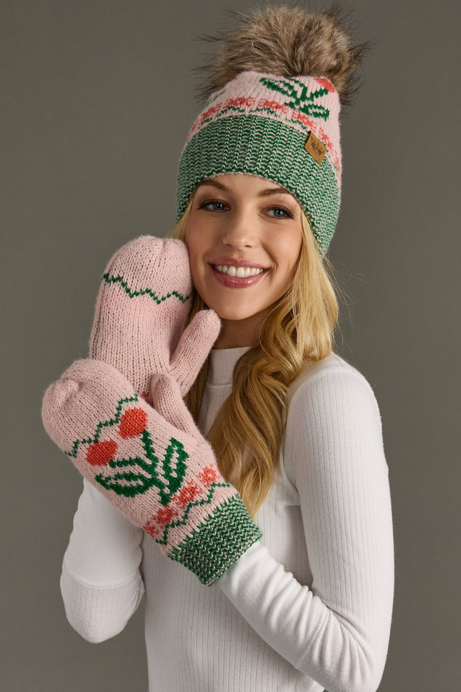 A smiling woman in a white sweater wears the Panache Apparel Collection Light Pink Cherry Pattern Pom Hat, a pink and green fleece-lined knit hat with a faux fur pom-pom, along with matching mittens decorated with flower patterns, as she holds her hands to her face against a gray background.
