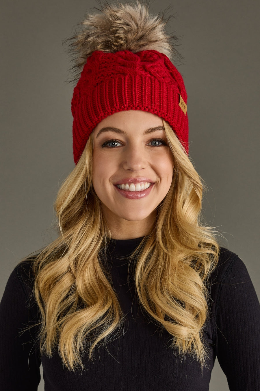 A woman with long blonde hair, wearing the Panache Apparel Collection Red Cable Knit Pom Hat and a black long-sleeve shirt, smiles against a plain gray background.