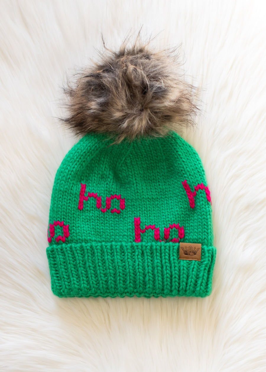 Green knit beanie with pink text and fur pom-pom on a white background