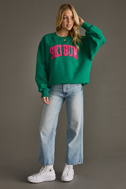 Person wearing a green 'Ski Bum' sweatshirt with blue jeans and white sneakers on a gray background