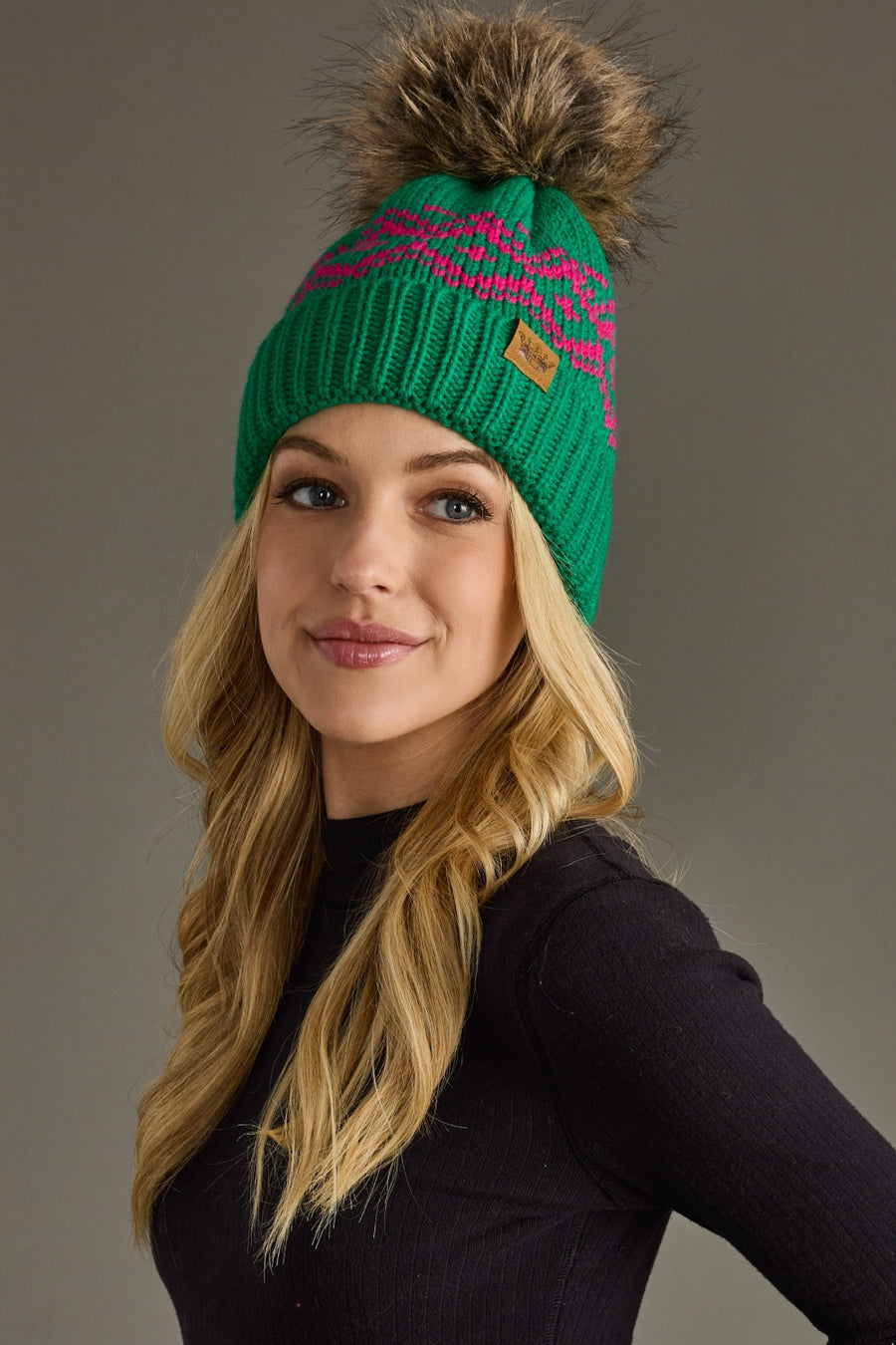 A woman with long blonde hair wears the cozy, fleece-lined After Hours Pom Hat by Panache Apparel Collection—a green knit hat with a pink pattern, large brown faux fur pom-pom, and black long-sleeve top—smiling against a gray background.