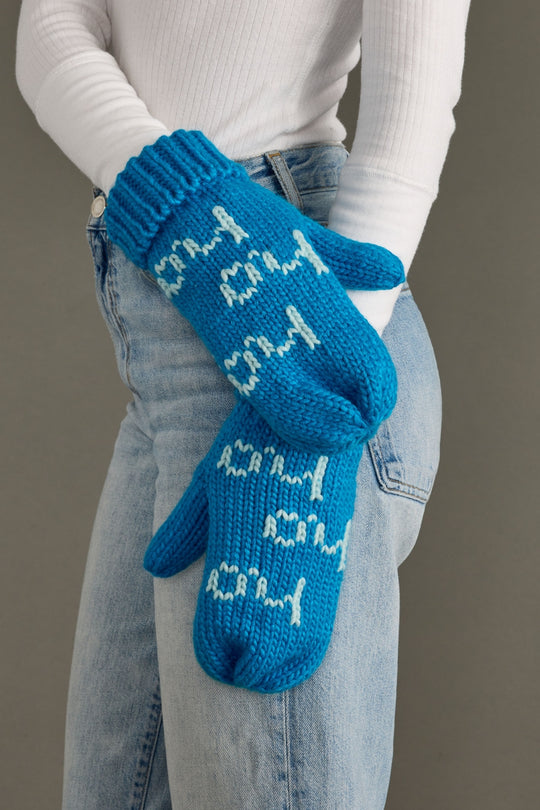 Blue mittens with text held by a person wearing jeans and a white shirt.
