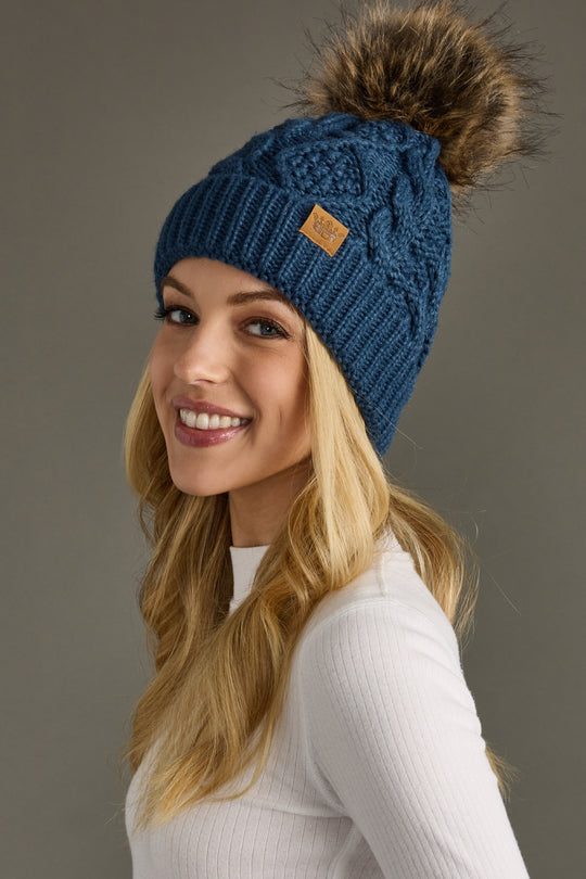 Woman wearing a blue knit beanie with a fur pom-pom on a gray background