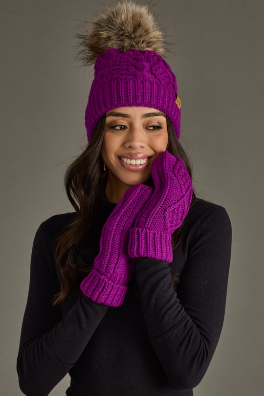 Woman wearing a purple knit hat with a fur pom-pom and matching mittens against a gray background